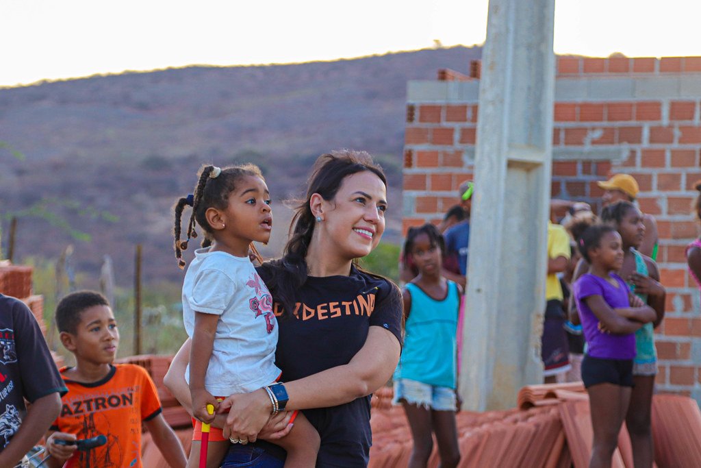 Talita Oliveira com uma menina no colo, cercada de crianças, ao lado de casas em construção